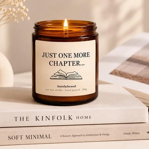 Könnte beinhalten: Eine brennende Sojawachskerze in einem braunen Glasgefäß mit cremefarbenem Etikett. Das Etikett zeigt den Text "JUST ONE MORE CHAPTER..." über einer Strichzeichnung eines offenen Buches sowie "Sandalwood", "soy wax candle", "hand-poured" und "200g".