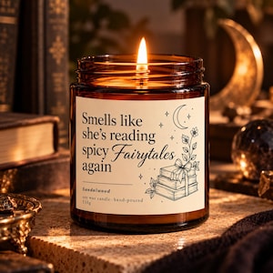 Könnte beinhalten: Eine brennende Sojawachskerze in einem bernsteinfarbenen Glasgefäß mit einem weißen Etikett, auf dem "Smells like she's reading spicy Fairytales again" und "Sandalwood" steht. Das Etikett zeigt eine Strichzeichnung eines Halbmonds und Bücher.