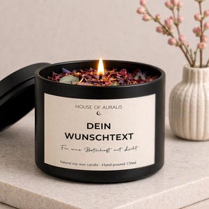 Könnte beinhalten: Eine brennende schwarze Kerze mit dem Text "DEIN WUNSCHTEXT" und "HOUSE OF AURALIS" auf dem Etikett. Die Kerze ist mit getrockneten Blumen gefüllt und steht auf einer Steinoberfläche. Eine kleine weiße Vase mit rosa Blumen befindet sich im Hintergrund.