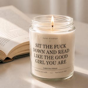 Könnte beinhalten: Eine brennende, transparente Glas-Kerze mit dem Text "SIT THE FUCK DOWN AND READ LIKE THE GOOD GIRL YOU ARE". Die Kerze ist mit "FAIRYTALE FOREST" und "Natural Soy wax candle" beschriftet. Im Hintergrund ein offenes Buch.