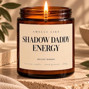 Könnte beinhalten: Eine brennende Sojawachskerze in einem bernsteinfarbenen Glasgefäß mit cremefarbenem Etikett. Der Text auf dem Etikett lautet: SMELLS LIKE SHADOW DADDY ENERGY, MYSTIC WOODS, soy wax candle, hand-poured, 200g. Die Kerze hat einen brennenden Docht.
