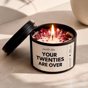 Könnte beinhalten: Eine schwarze Metalldose mit einer Kerze und einem weißen Etikett mit der Aufschrift "smells like YOUR TWENTIES ARE OVER" und "Scent: Vanilla & Oud". Die Kerze brennt, mit einer sichtbaren Flamme und getrockneten Blütenblättern auf dem Wachs.