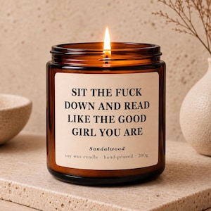 Könnte beinhalten: Eine brennende Sojawachskerze in einem braunen Glasgefäß mit einem cremefarbenen Etikett. Der Text auf dem Etikett lautet: SIT THE FUCK DOWN AND READ LIKE THE GOOD GIRL YOU ARE. Darunter steht Sandalwood, soy wax candle, hand-poured, 200g.