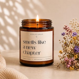 Könnte beinhalten: Eine brennende Kerze in einem braunen Glas mit dem Text "smells like a new chapter". Die Kerze steht neben einem Strauß getrockneter Blumen. Das Glas ist mit "House of Auralis" beschriftet.