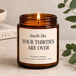 Könnte beinhalten: Eine brennende Sojawachskerze in einem bernsteinfarbenen Glasgefäß mit cremefarbenem Etikett. Der Text auf dem Etikett lautet: Smells like YOUR THIRTIES ARE OVER, Himalaya Cashmere, soy wax candle - hand-poured - 200 g. Die Kerze hat einen Docht.