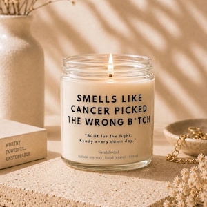 Könnte beinhalten: Eine Kerze im klaren Glasgefäß mit weißem Wachs und brennendem Docht. Das Etikett trägt die Aufschrift: SMELLS LIKE CANCER PICKED THE WRONG B*TCH, Built for the fight. Ready every damn day., Sandalwood, natural soy wax - hand poured - 180 ml.