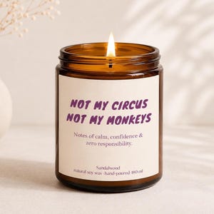 Könnte beinhalten: Eine brennende Kerze in einem bernsteinfarbenen Glas mit einem weißen Etikett, das den violetten Text "NOT MY CIRCUS NOT MY MONKEYS" sowie "Notes of calm, confidence & zero responsibility." und "Sandalwood natural soy wax hand-poured 180 ml" zeigt.