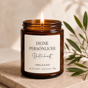 Könnte beinhalten: Eine brennende Sojawachskerze in einem braunen Glasgefäß mit einem weißen Etikett, das den Text DEINE PERSÖNLICHE Botschaft, VANILLA & OUD sowie soy wax candle · hand-poured · 200 g trägt. Die Kerze steht auf einem Stein neben einer Pflanze.