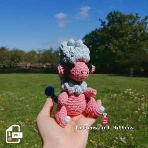 Puede incluir: Un juguete amigurumi de crochet rosa y azul que se asemeja a una oveja. El juguete tiene una cola de pompón azul y está sostenido en la mano de una persona. El fondo es un campo de hierba verde con flores.
