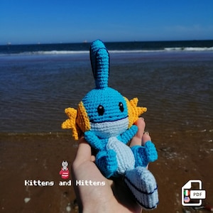 Pode incluir: Um personagem Pokemon de crochê azul e amarelo, com um rosto branco e olhos pretos, está sendo segurado em uma mão. O personagem está em uma praia com o oceano ao fundo. O texto "Mittens and Kittens" é visível na imagem.