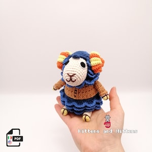 May include: Crocheted amigurumi toy of a sheep with blue and brown clothing. The sheep has yellow and orange curly horns. The toy is being held in a hand.