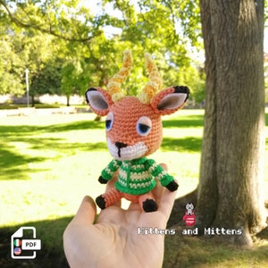 Könnte beinhalten: Gehäkelte Gazellenfigur mit orangefarbenen, weißen, grünen und schwarzen Details. Die Gazelle hat große blaue Augen, gelbe und orangefarbene Hörner und trägt einen grün-weiß gestreiften Pullover. Der Text "Kittens and Mittens" ist sichtbar.