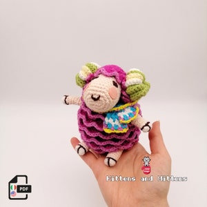 May include: A crocheted pink sheep with green and yellow striped horns, wearing a blue and white striped scarf. The sheep is sitting on a hand.