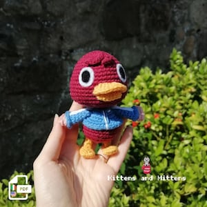 May include: Crocheted amigurumi toy of a red duck wearing a blue and white jacket. The duck has a yellow beak and large black eyes. The toy is being held in a person's hand. The image includes the text "Kittens and Mittens".