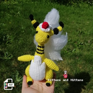 May include: A crocheted yellow and black bee Pokemon plush toy with a white mane and a red and white hat. The toy is being held in a hand. The text " Kittens and Mittens" is visible in the image.