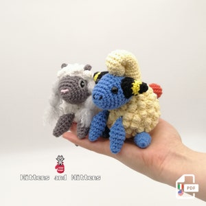May include: Two crocheted amigurumi plushies, one is a light blue and yellow sheep with black markings and the other is a gray sheep with white fur. The plushies are sitting in a person's hand. The text "Kittens and Mittens" is below the plushies.