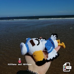 Peut inclure: Un jouet en crochet en forme de mouette, habillé d'un costume de marin bleu et blanc. Le jouet est posé sur une main avec l'océan en arrière-plan.