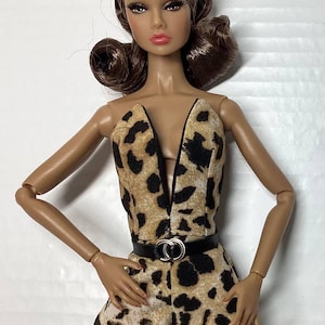 May include: A fashion doll with brown hair styled in curls, wearing a sleeveless jumpsuit featuring a tan and black leopard print pattern. The jumpsuit has a deep V-neckline and is cinched at the waist with a thin black belt featuring a silver buckle.