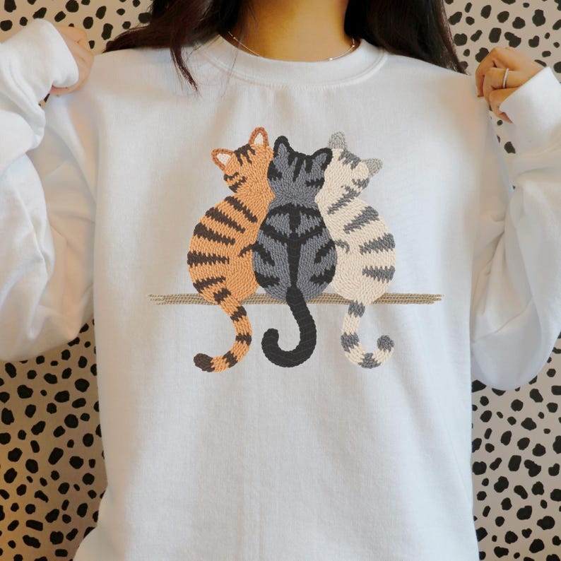 K&ouml;nnte beinhalten: Wei&szlig;es Sweatshirt mit einem gestickten Design von drei Katzen. Die Katzen sind in den Farbt&ouml;nen Orange, Grau und Creme gehalten und sitzen zusammen auf einer braunen Linie. Das Sweatshirt hat lange &Auml;rmel und einen Rundhalsausschnitt.