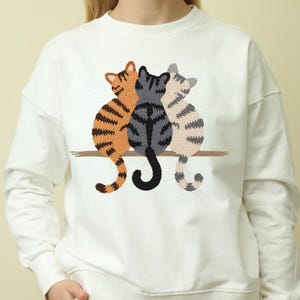 K&ouml;nnte beinhalten: Wei&szlig;es Sweatshirt mit einer Stickerei von drei Katzen, die auf einem Ast sitzen. Die Katzen sind in Orange, Grau und Beige gehalten und haben Streifenmuster. Das Sweatshirt hat einen Rundhalsausschnitt und lange &Auml;rmel.