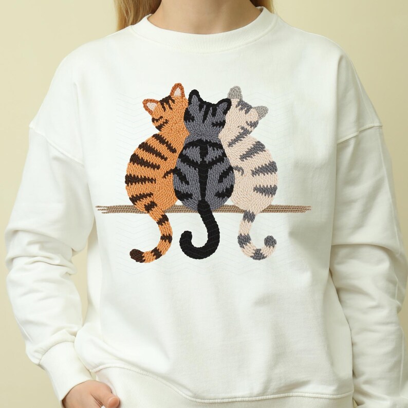 K&ouml;nnte beinhalten: Wei&szlig;es Sweatshirt mit einer Stickerei von drei Katzen, die auf einem Ast sitzen. Die Katzen sind in Orange, Grau und Beige gehalten und haben Streifenmuster. Das Sweatshirt hat einen Rundhalsausschnitt und lange &Auml;rmel.