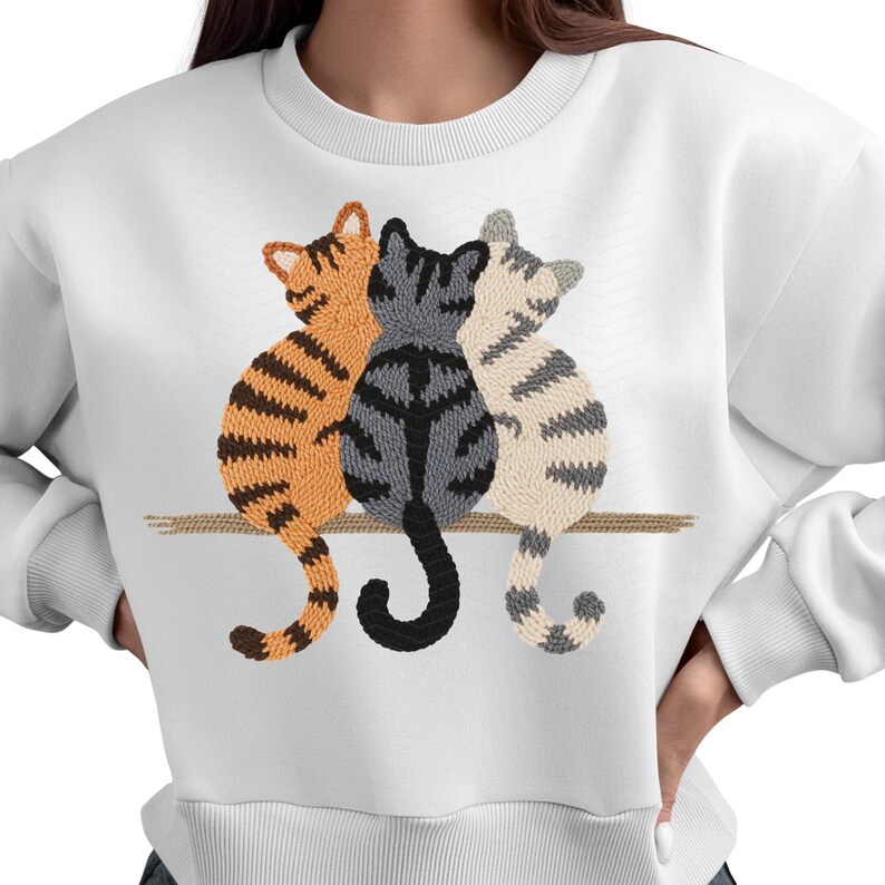 K&ouml;nnte beinhalten: Wei&szlig;es Sweatshirt mit einer Stickerei von drei Katzen, die nebeneinander sitzen. Die Katzen sind orange, grau und cremefarben, mit gestreiften Mustern. Das Design befindet sich auf einer horizontalen braunen Linie.