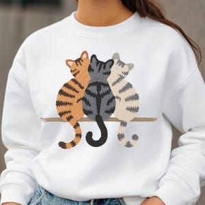 K&ouml;nnte beinhalten: Wei&szlig;es Sweatshirt mit einem gestickten Design von drei Katzen, die nebeneinander auf einem braunen Ast sitzen. Die Katzen sind in Orange-, Grau- und Cremet&ouml;nen gehalten und weisen detaillierte Muster auf. Das Sweatshirt ist ein l&auml;ssiger, bequemer Stil.