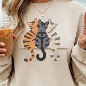 May include: Beige sweatshirt featuring an embroidered design of three cats. The cats are in shades of orange, gray, and white, with detailed striped patterns. The cats are sitting together on a brown line.