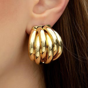 May include: Close-up of a gold-colored, chunky hoop earring. The earring has a ribbed design with multiple curved segments, creating a bold, textured appearance. The earring is worn on the ear, showcasing its polished, reflective surface.