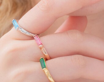 May include: Three colorful rings with rectangular gemstones. One ring has a light blue gemstone, another has a pink gemstone, and the third has a green gemstone. The rings are on a hand with a light skin tone.