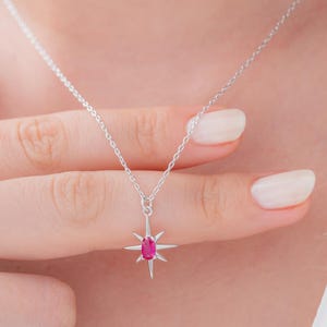 May include: A silver necklace with a starburst pendant featuring a vibrant pink gemstone at its center. The pendant is suspended from a delicate chain. The necklace is displayed on a person's neck.