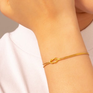 May include: A gold bracelet with a knot design. The bracelet is made of a thin, braided chain.