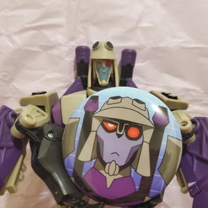 May include: A purple and tan Transformers action figure holding a round button with a cartoon image of a robot. The button has a blue background and a purple robot graphic.