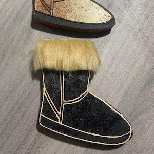 May include: Two decorative wooden boot ornaments. The top boot is gold with brown fur trim. The bottom boot is black with light brown fur trim.