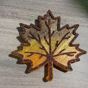 May include: A glittery gold and brown maple leaf ornament with a red glitter outline. The leaf is made of a textured material and has a three-dimensional design.