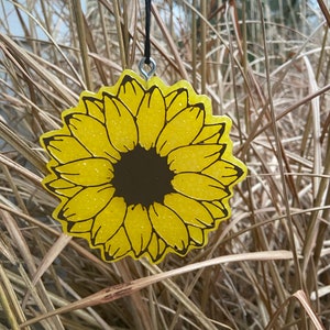 May include: A yellow sunflower with a brown center, hanging from a black string. The sunflower is made of a glittery material.
