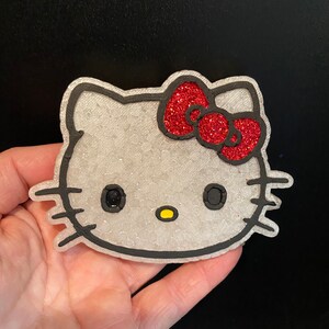 May include: A white Hello Kitty character with black outlines and a red glitter bow. The character has black eyes and a yellow nose.