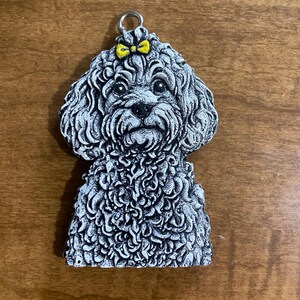 May include: A gray, textured dog pendant featuring a curly-haired poodle with a small, bright yellow bow on its head. The pendant has a silver-toned metal jump ring attached to the top, allowing it to be used as a charm or decorative ornament.