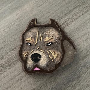 May include: A detailed, brown and tan dog head sculpture with a stern expression. The sculpture features a black nose, white eyes, and a pink tongue. The dog's head is outlined in a darker brown, and the background is a light gray wood grain.