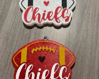 May include: Two white and red football-shaped ornaments with the word "Chiefs" in white lettering. The ornaments have a black and gold football design with a small red heart on the white ornament and a black heart on the red ornament.