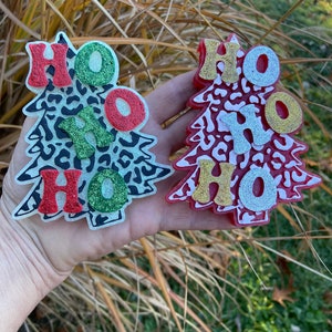May include: Two Christmas tree ornaments with a leopard print design. The ornaments are decorated with the word "HO HO HO" in red, green, gold, and silver glitter.