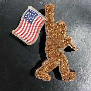 May include: A gold glitter silhouette of a person holding a small American flag. The flag is red, white, and blue.