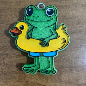 May include: A green frog wearing blue swim shorts and a yellow duck-shaped inflatable ring. The frog has large black eyes and is smiling. The image is a cartoon-style illustration.