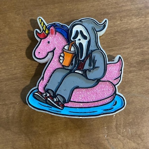 May include: A quirky sticker showing a Ghostface character from the Scream movies on a pink glitter unicorn float in a blue pool. The character is drinking from a cup and wearing grey clothes and red and black trainers.