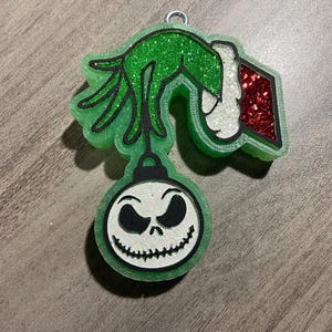 May include: Green glitter Christmas ornament featuring the Grinch's hand holding a red and white Santa hat with a green glitter ornament of Jack Skellington's face hanging from it.