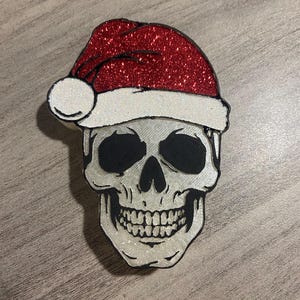 May include: A white skull with a black outline wearing a red glitter Santa hat with a white pom-pom.