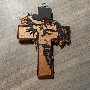 May include: A wooden cross pendant with a detailed depiction of Jesus's face and crown of thorns. The cross is brown with black accents and a metal loop for hanging. The design is carved into the wood.