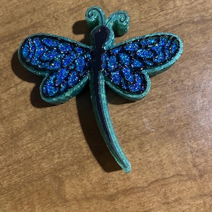 May include: A dragonfly-shaped resin ornament. The wings are a vibrant blue with glitter, outlined in black, and the body is a dark green. The dragonfly has a curved tail and is set against a wooden surface.