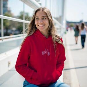 May include: A red hoodie with a white scooter design on the front. The hoodie has a drawstring hood and a front pocket. The person is wearing blue jeans. The background is a bridge with people walking.
