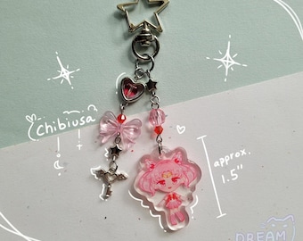 Può includere: Un portachiavi in acrilico rosa e trasparente con un personaggio dei cartoni animati, un fiocco e un ciondolo a forma di chiave. Il portachiavi include una chiusura a forma di stella ed è lungo circa 3,8 cm. Il testo "Chibiusa" è scritto a sinistra.